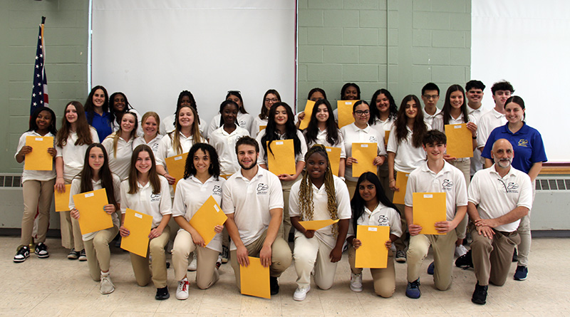 A group of 28 high school students, all wearing white polo shirts and tan pants, along with three adults, pose together. The students are all holding large envelopes.