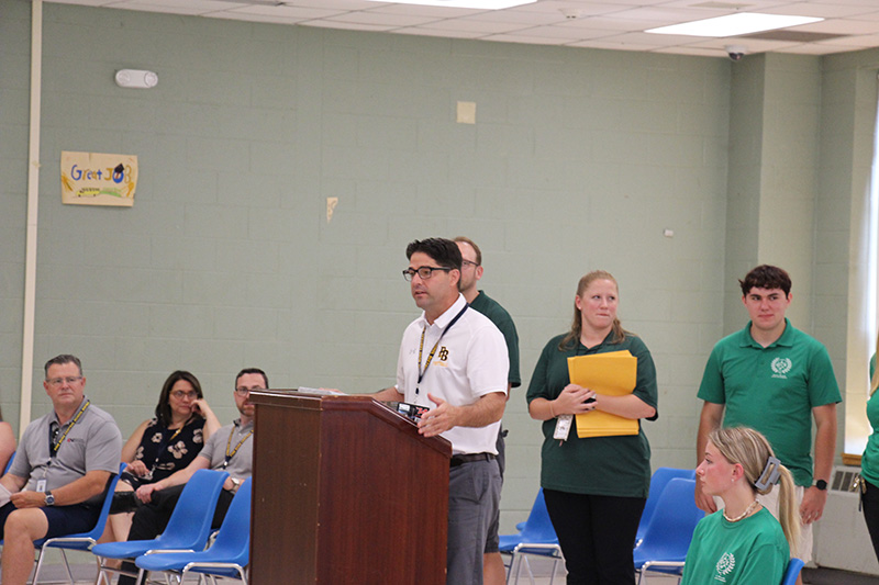 A man in a white short-sleeve polo  shirt stands at a podium speaking. There are a few people behind him.