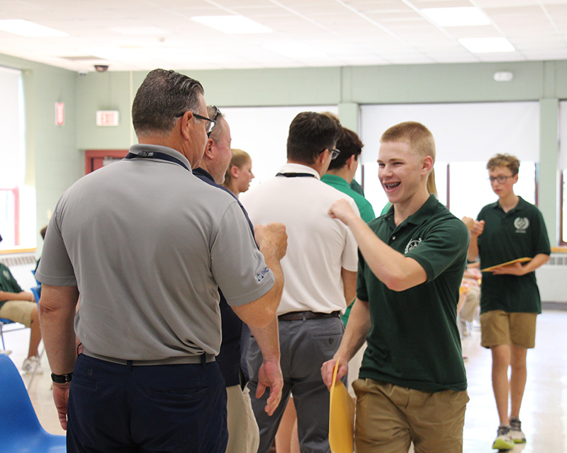 A high school kid with short blonde hair wearing a green shirt fist bumps a man.