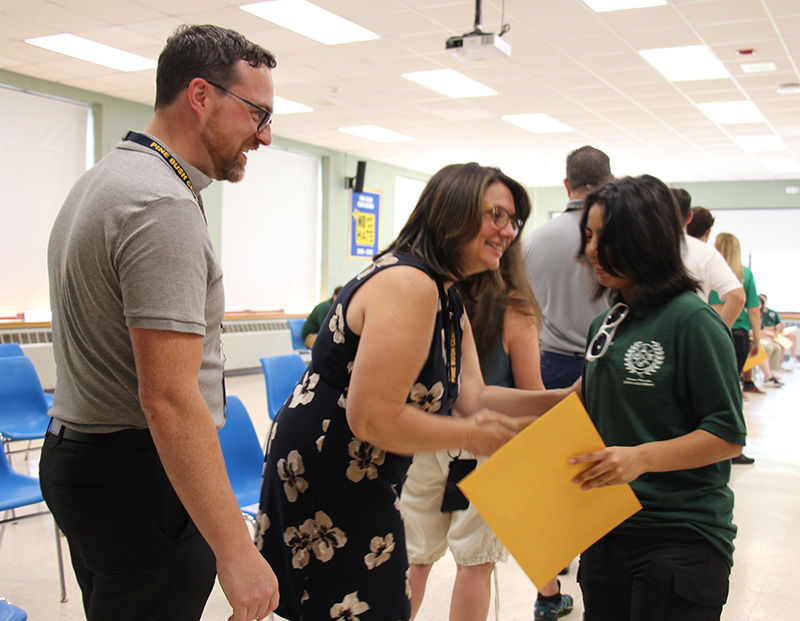 A high school student wearing a green shirt shakes hands with a woman who is bending down to congratulate her. A man in a short-sleeve shirt, wearing glasses, smiles.