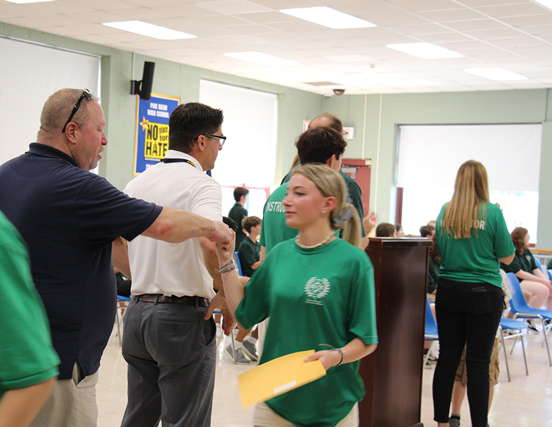 A young woman with long blonde hair wearing a green shirt fist bumps a man in a blue shirt.