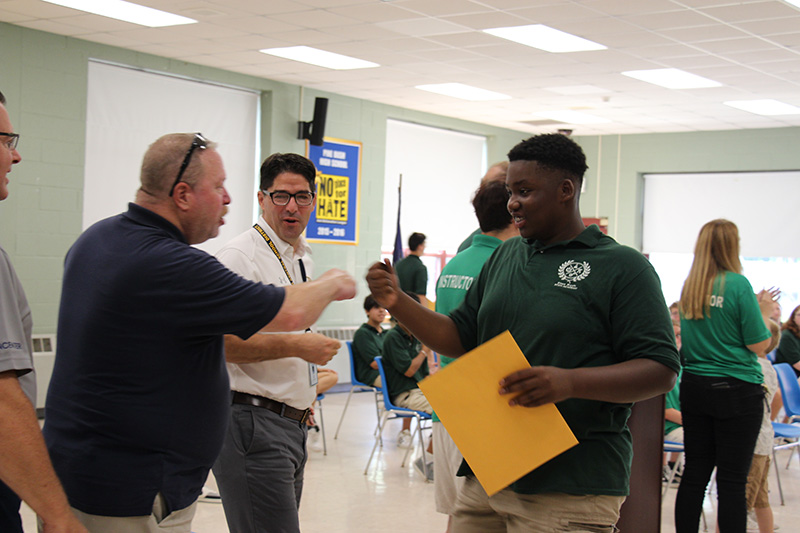 A high school boy wearing a green shirt fist bumps a man in a blue shirt while  man in a white shirt turns and smiles.