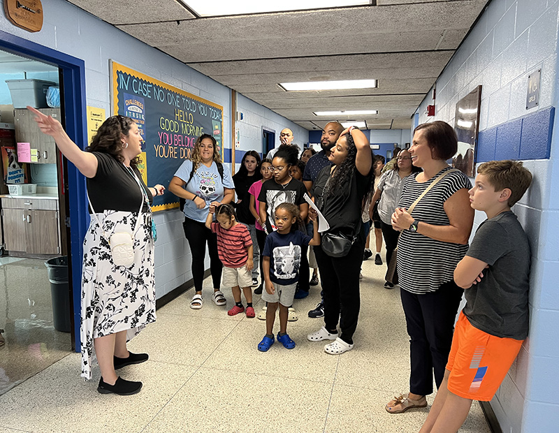 A hallways in a school with lots of adults and children. One adult on the left is pointing down the hall as she gives a tour.