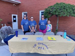 Three adults and two children stand behind a table with a tan table cloth that says EJR Elementary PTA.