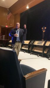 A man in a dress shirt and jacket with Khaki pants walks in the front of an auditorium talking.