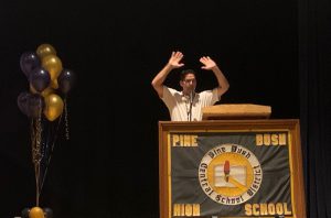 A man wearing a white polo shirt stands at a podium, with a sign in front that says Pine Bush High School, holds his hand up in the air. There are balloons on the let side of the stage.