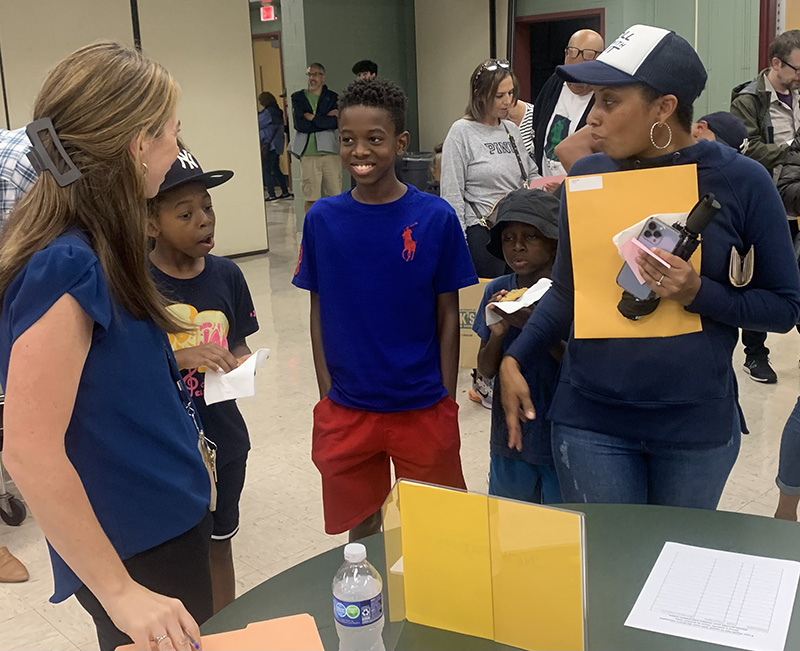 Two young men stand and smile as they talk to two women at an activities fair in a high school.