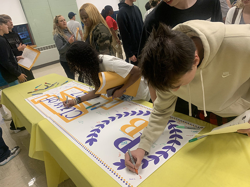 Two incoming freshmen lean over a table with a gold table cloth and sign a banner that says PB class of 2027.