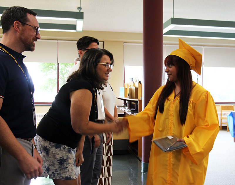 A young woman in a gold cap and gown with long brown hair shakes the hand of a woman  while holding her diploma in the other.