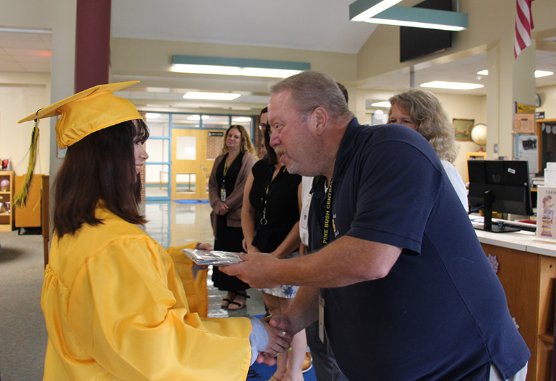 A young woman in a gold cap and gown with shoulder-length brown hair shakes the hand of a man  while holding her diploma in the other.