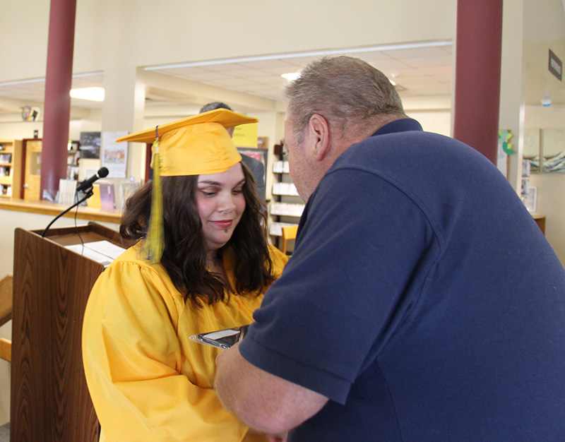 A young woman in a gold cap and gown with long brown hair shakes the hand of man  while receiving her diploma in the other.