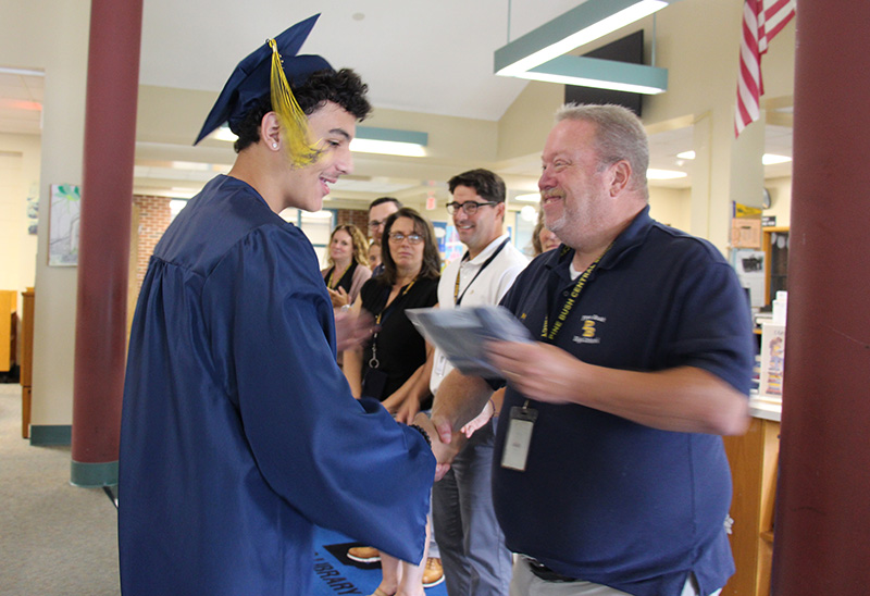 A young man in a blue cap and gown shakes hands with a man as he receives his high school diploma. Both are smiling as are the others who are looking on.