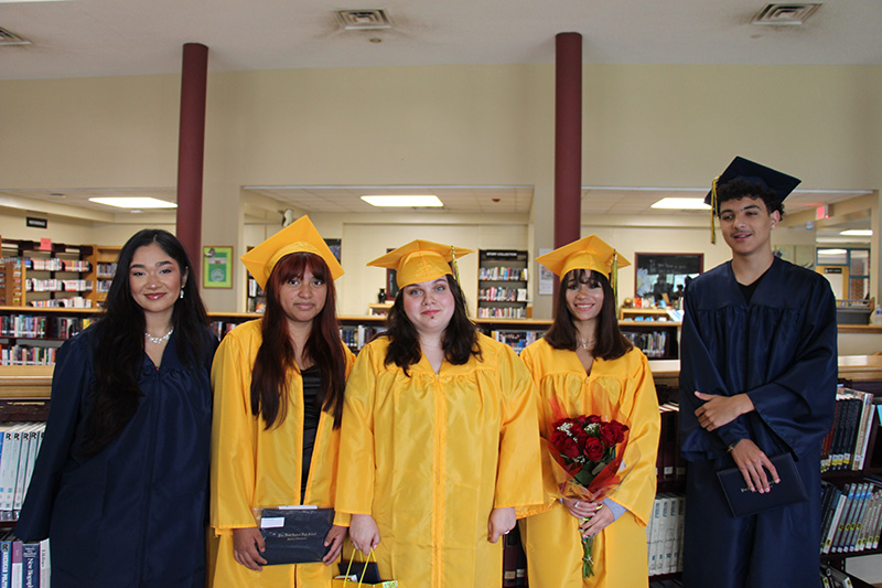 Five young people - four young women and one young man in blue or gold caps and gowns - stand together smiling. One girl is holding flowers and another her diploma.