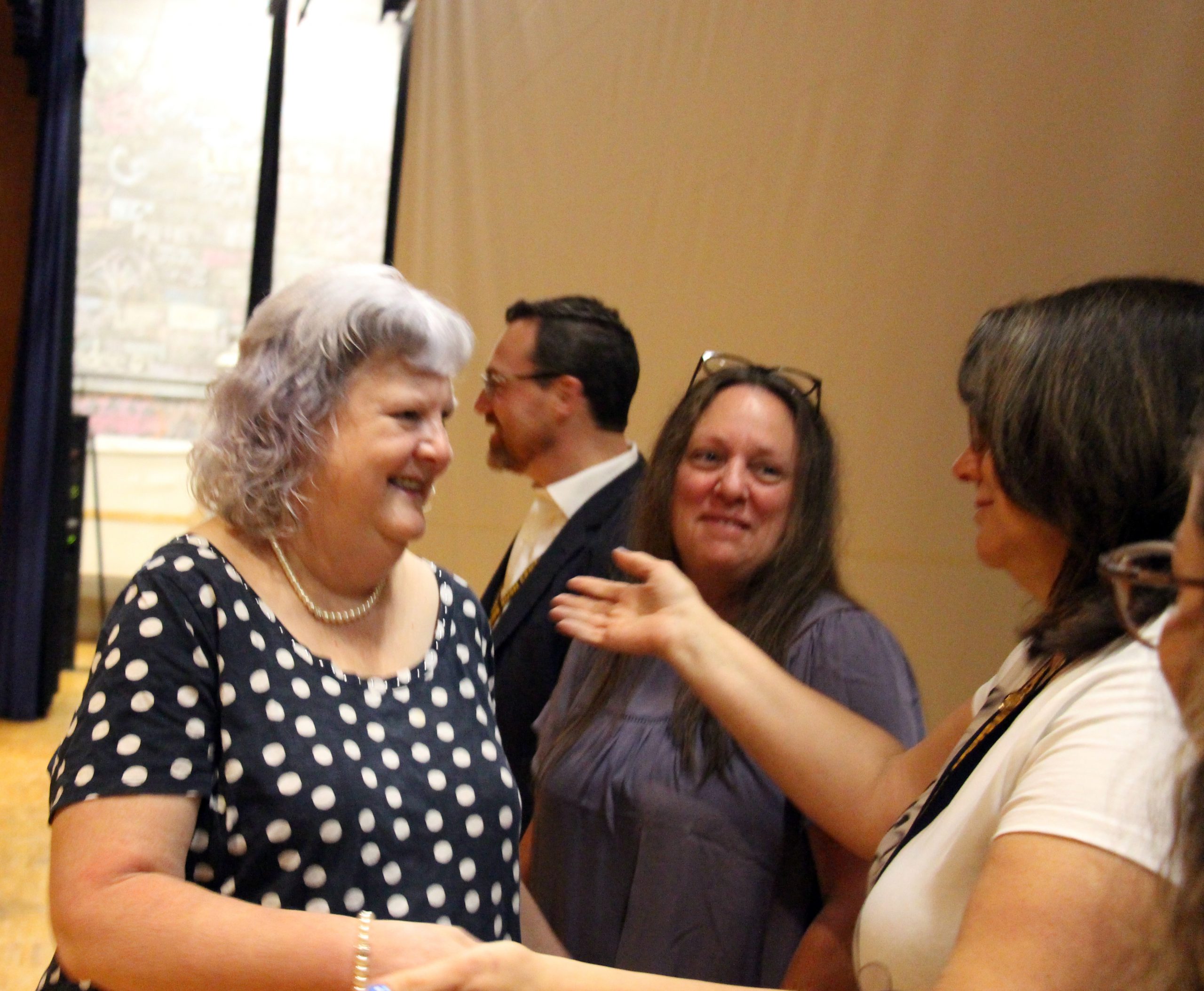 A woman with gray short hair, wearing a blue and white polka dot shirt, smiles as she goes to hug a woman whose arms are open.