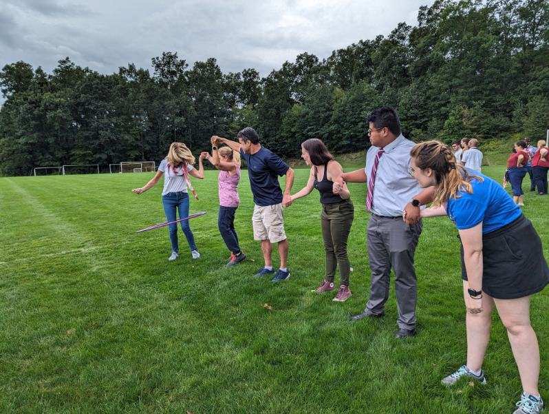 A group of seven adults in a straight line holding hands. The woman at the end has a hula hoop around her body.