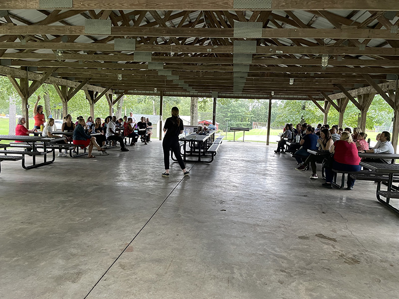 A woman stands before tables filled with adults under a wooden canopy.