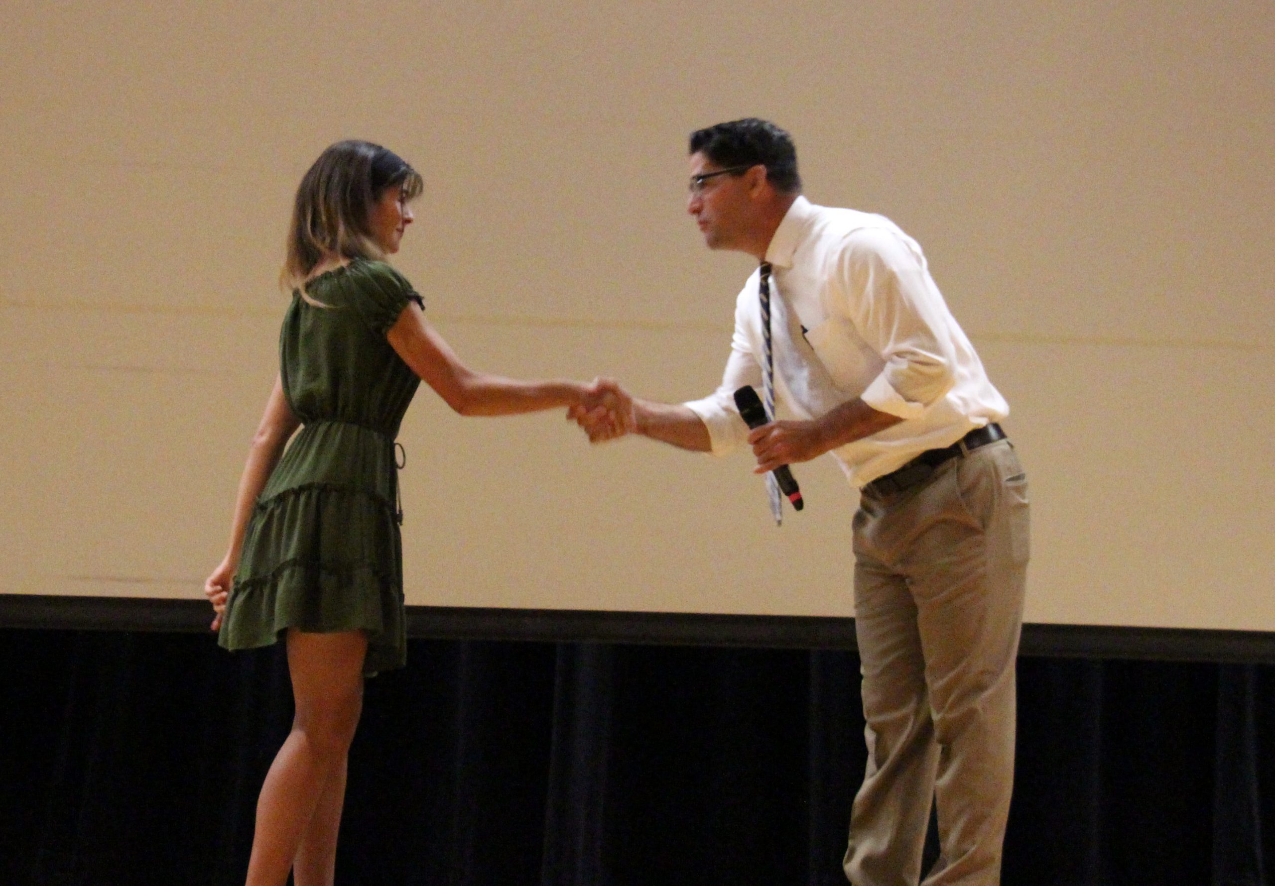 A man in a white shirt and tie leans in and shakes the hand of a young woman in a green dress.