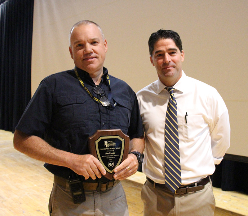 Two men stand together on a stage. The man on the left is wearing a navy blue shirt and is smiling as he holds a plaque. the man on the right has dark hair and is wearing a white shirt and blue and gold striped tie. He is smiling as well.