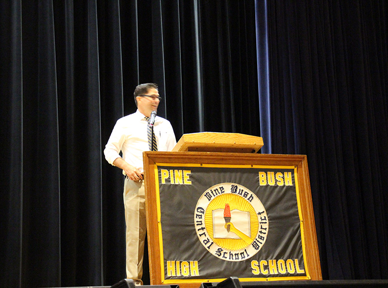 A large sign that says Pine Bush High School with a logo in the center is in front of a dais where a man with short dark hair is speaking.