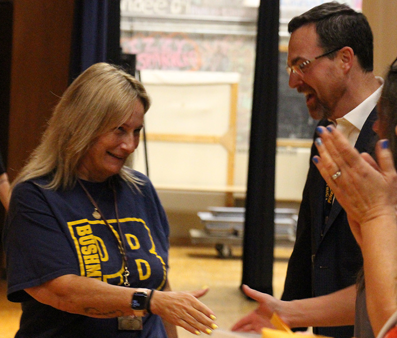 A woman with long blonde hair wearing a blue shirt with a large PB in gold on it, goes to shake hands with a woman. Another woman claps.