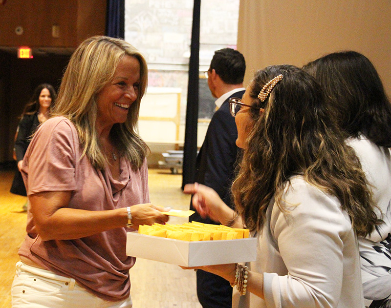 A woman with long blonde hair smiles as she accepts a small envelop from a woman on the right.