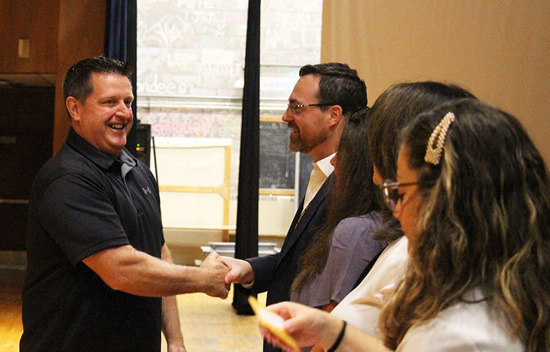 A man with short dark hair wearing a dark shirt, shakes hands with a man with short dark hair and glasses. Both are smiling.