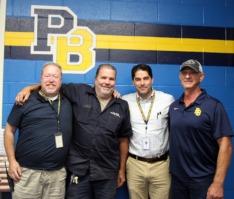 Four men stand arm-in-arm and smile. Behind them on the wall is a painted PB in blue and gold.