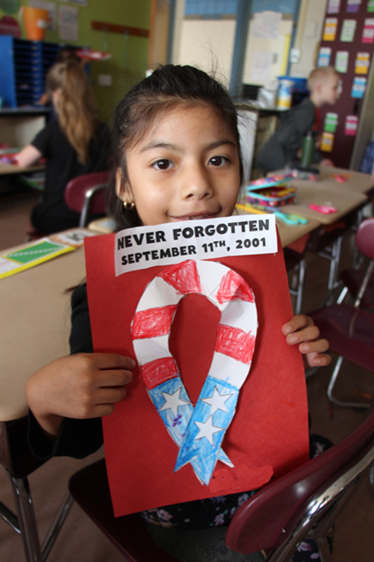 A girl smiles and holds a red piece of paper with a red white and blue ribbon on it. It says Never Forgotten September 11, 2001