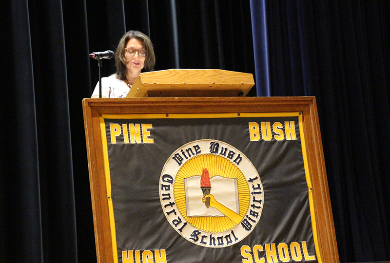 A large sign that says Pine Bush High School with a logo in the center is in front of a dais where a woman with shoulder-length dark hair is speaking.