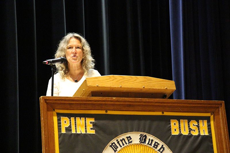 A large sign that says Pine Bush High School with a logo in the center is in front of a dais where a woman with shoulder-length blonde hair is speaking.