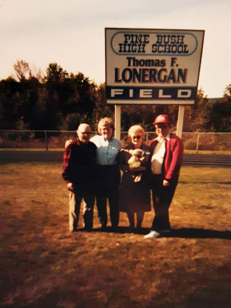 An old photo. Four adults standing in front of a sign that says Pine Bush Central School District Thomas F. Lonergan Field.