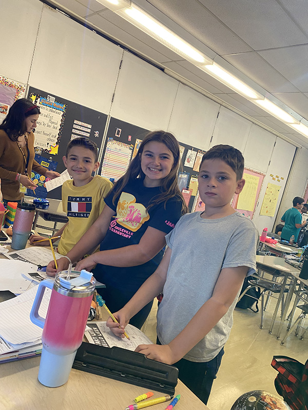 Three fifth-grade students stand at a table and smile as they write in their notebooks.