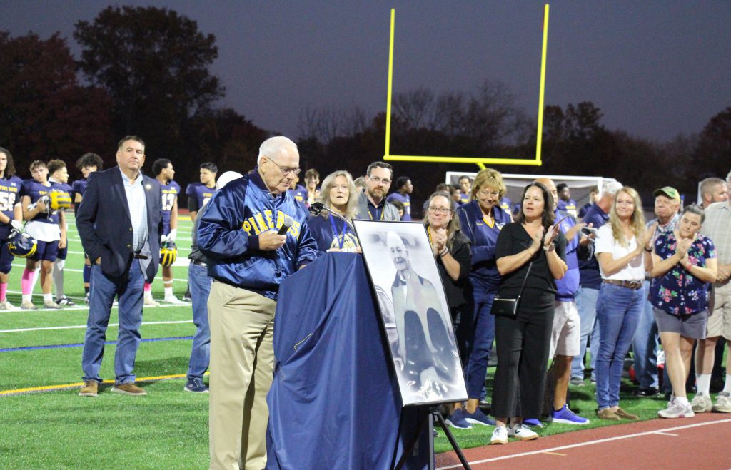 A group of people stand and listen as a man stands behind a podium covered in a blue cloth. In front of him is a large black and white photo of a man.