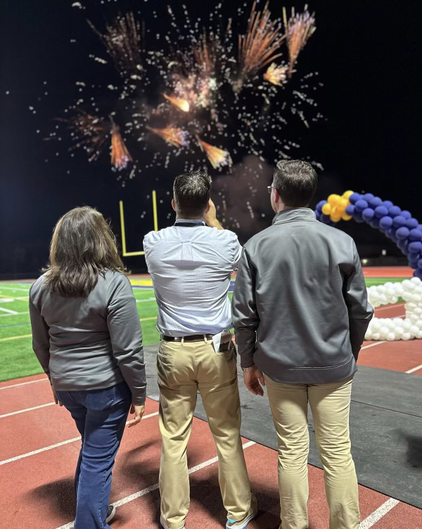 A woman and two men stand on a track and look up to see fireworks burstin above a football field.