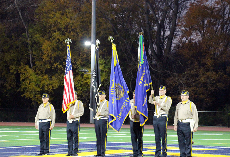 Six men in uniform hold four flags, including an American flag. They are standing in the center of a playing field.