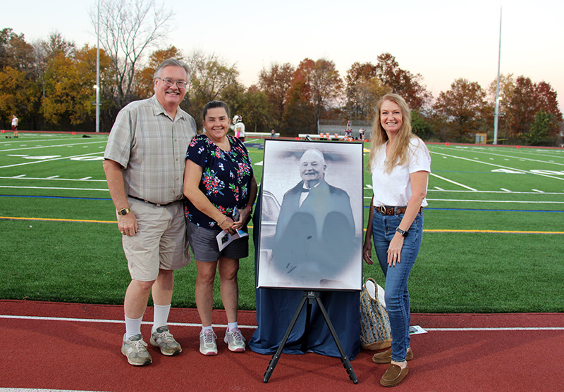 A man at left, a woman next, and a woman with long blonde hair on the right, flank a large poster showing an older man from the 1960s. Behind them is a football field.