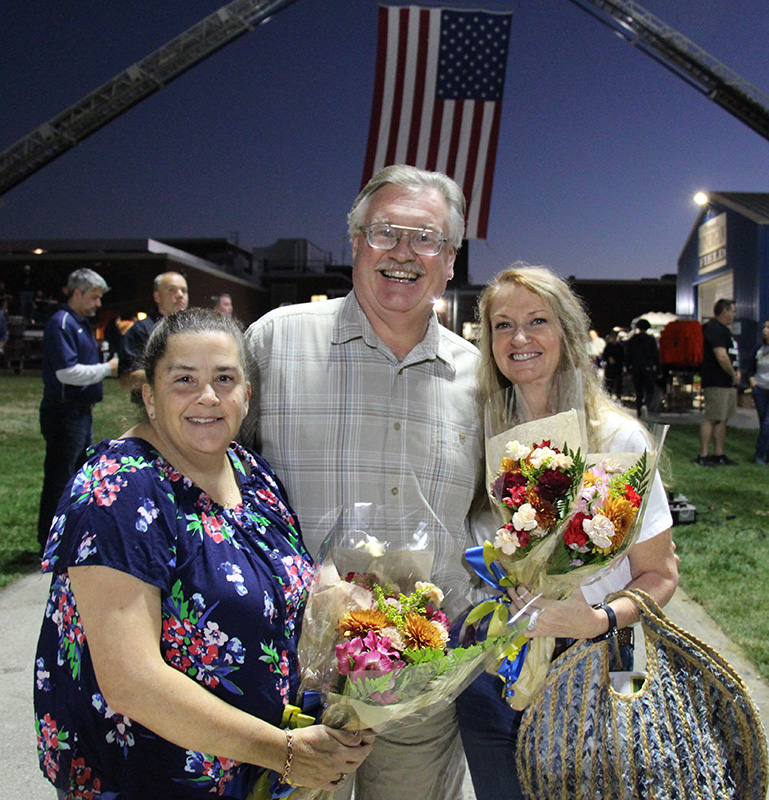 A woman, man in the center, and another woman all smile holding flowers. There is a large American flag behind them and it is getting dark out.