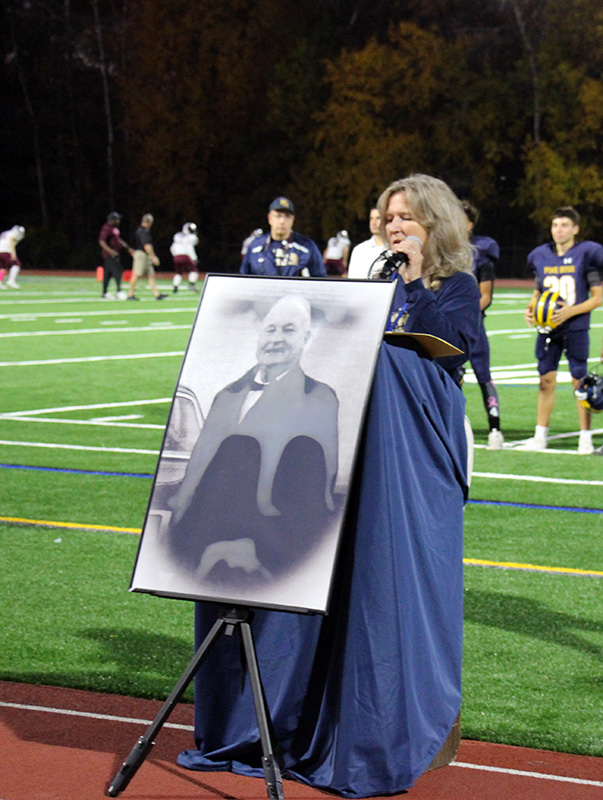 A woman with  shoulder-length blonde hair stands behind a podium draped in blue and speaks to a crowd. In front of her is a large poster of a man from the the 1960s. behind her is a playing field.