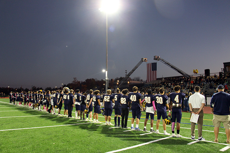 A line of football players, wearing navy blue unifoems with white numbers on their backs, stand at dusk on a field. There is a huge American flag in the back ground.