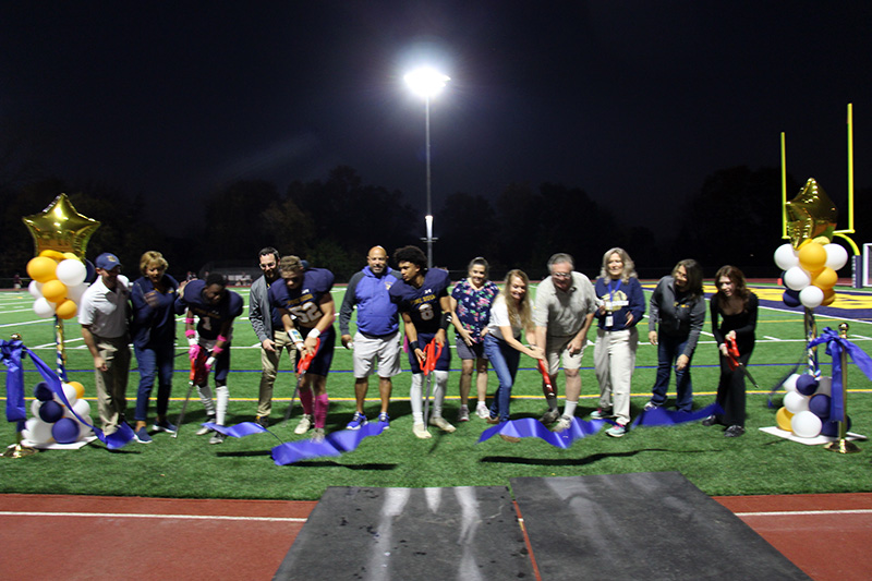 A group of four high school students and two adults hold oversize scissors after cutting a large blue ribbon on a field.