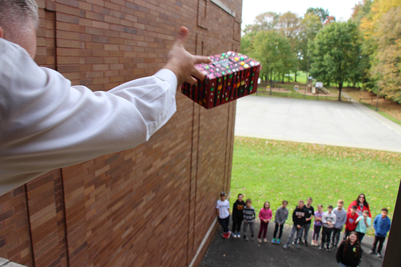 From the inside of the building, this is a picture of a man hanging out the window and tossing a box. Below, on the perimeter, are kids looking up and watching.