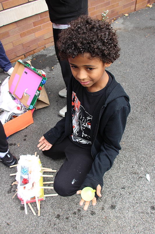 A boy with dark curly hair, wearing a black shirt and pants, holds a small water balloon in his hand and smiles.