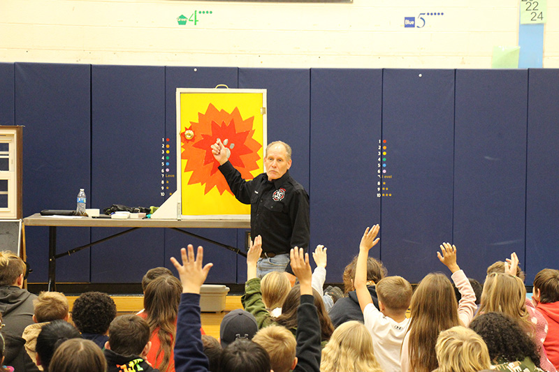 A man stands in front of a group of elementary students pointing to a photo of a fire. Many of the kids have their hands up.