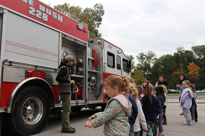 A group of elementary students line up listening to a man talk. Behind him is a red and white fire truck.