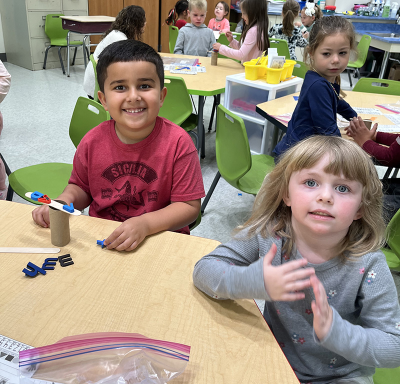 A kindergarten boy and girl sit at a table and smile with a little tree they built.