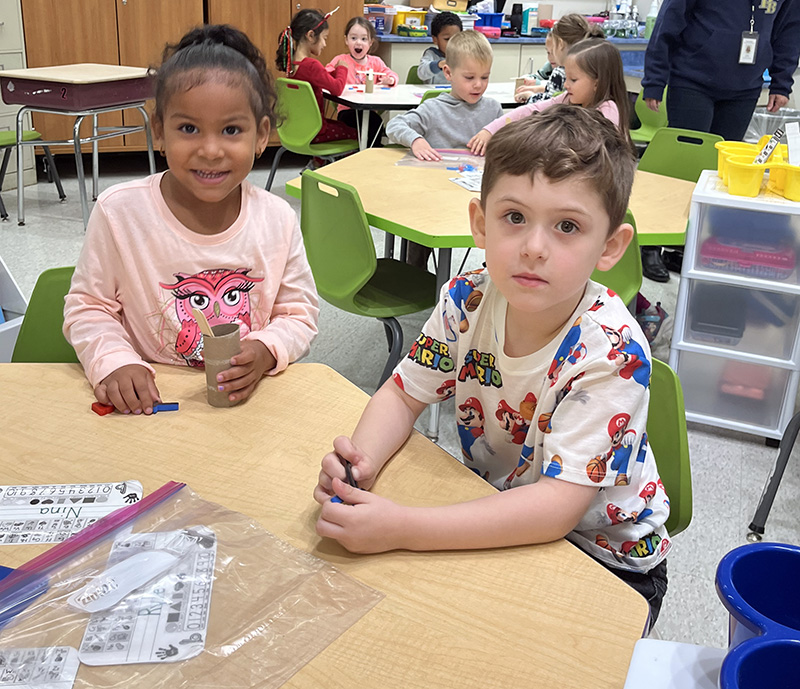 A girl on the left and a boy on the right sit at a table as they built a little tree.