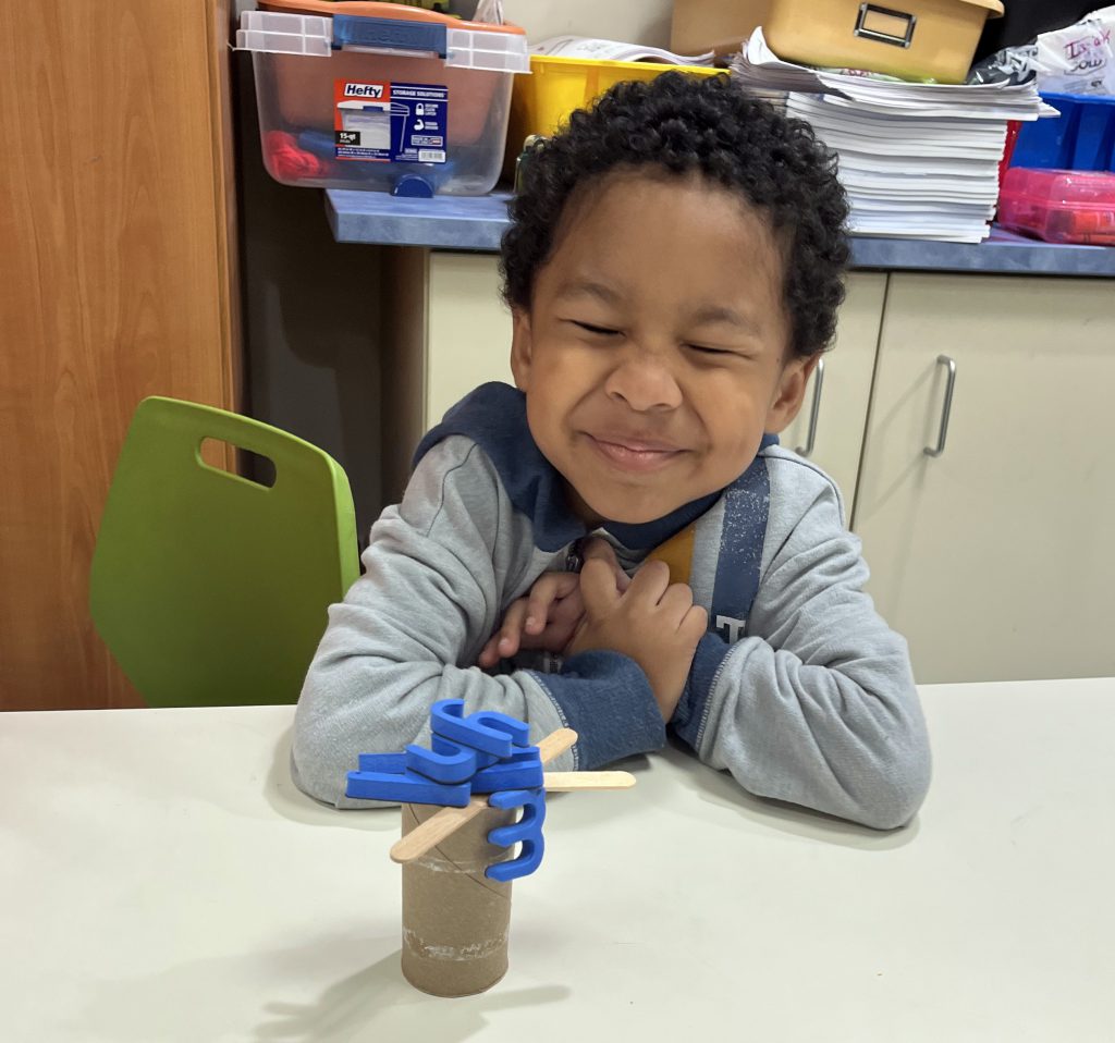 A kindergarten boy closes his eyes as he squishes his face smiling. His little tree, made of a paper roll and popsicle sticks is on the table. There are blue letters on the branches.