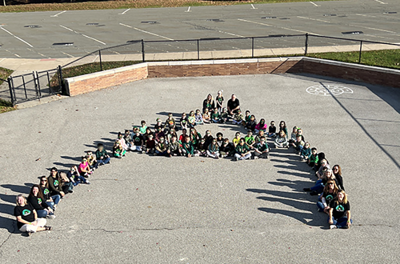 About 60 children and 10 adults form a large A on a courtyard with the photo taken from above.