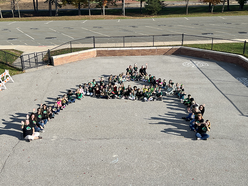 About 60 children and 10 adults form a large A on a courtyard with the photo taken from above. They are all waving.