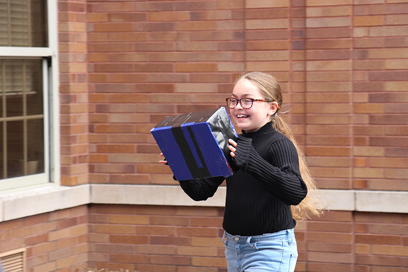 A girl with long blonde hair in a ponytail, wearing glasses and a dark shirt, smiles broadly as she picks up a box wrapped in blue tape.